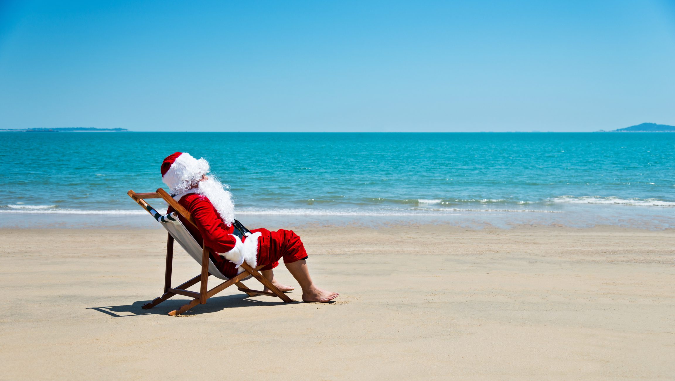 Santa Claus sitting on a beach chair on a sandy beach with the ocean in the background.