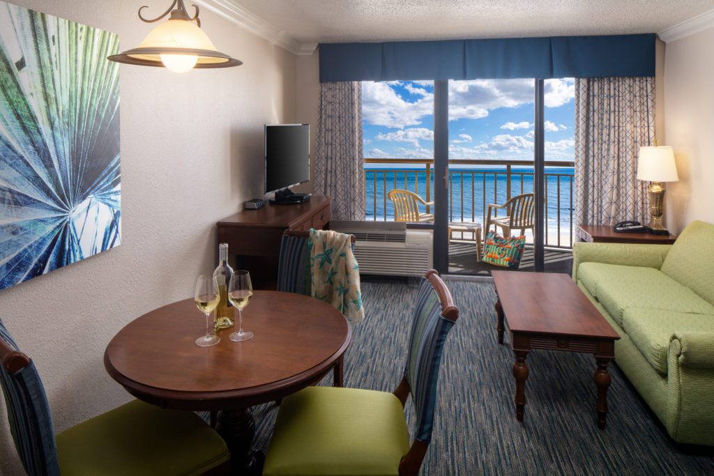 Holiday Inn suite living room with dining table set with wine glasses, green sofa, flat-screen TV, and oceanfront balcony