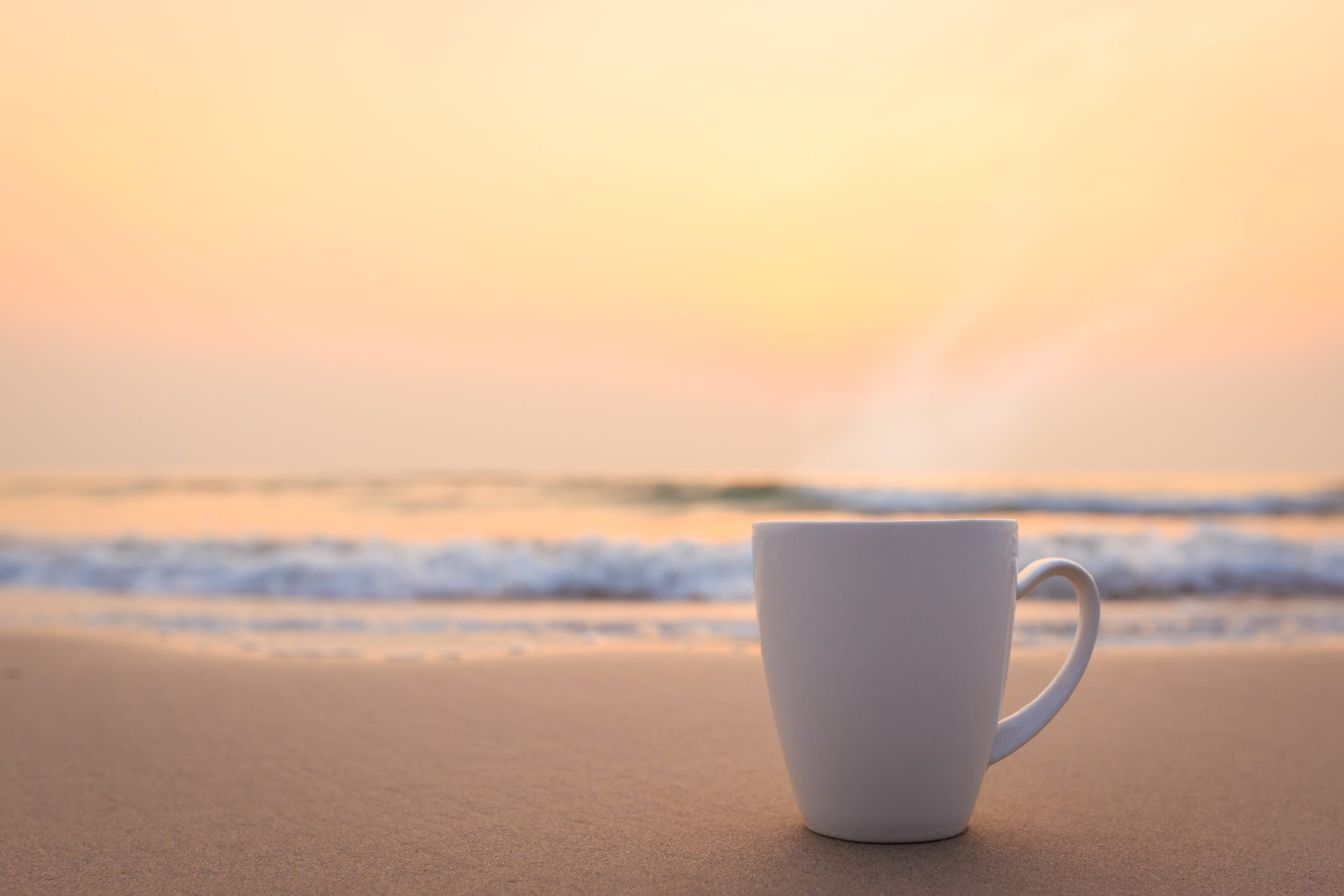 Close up white coffee cup on sand beach and view of sunset or sunrise background