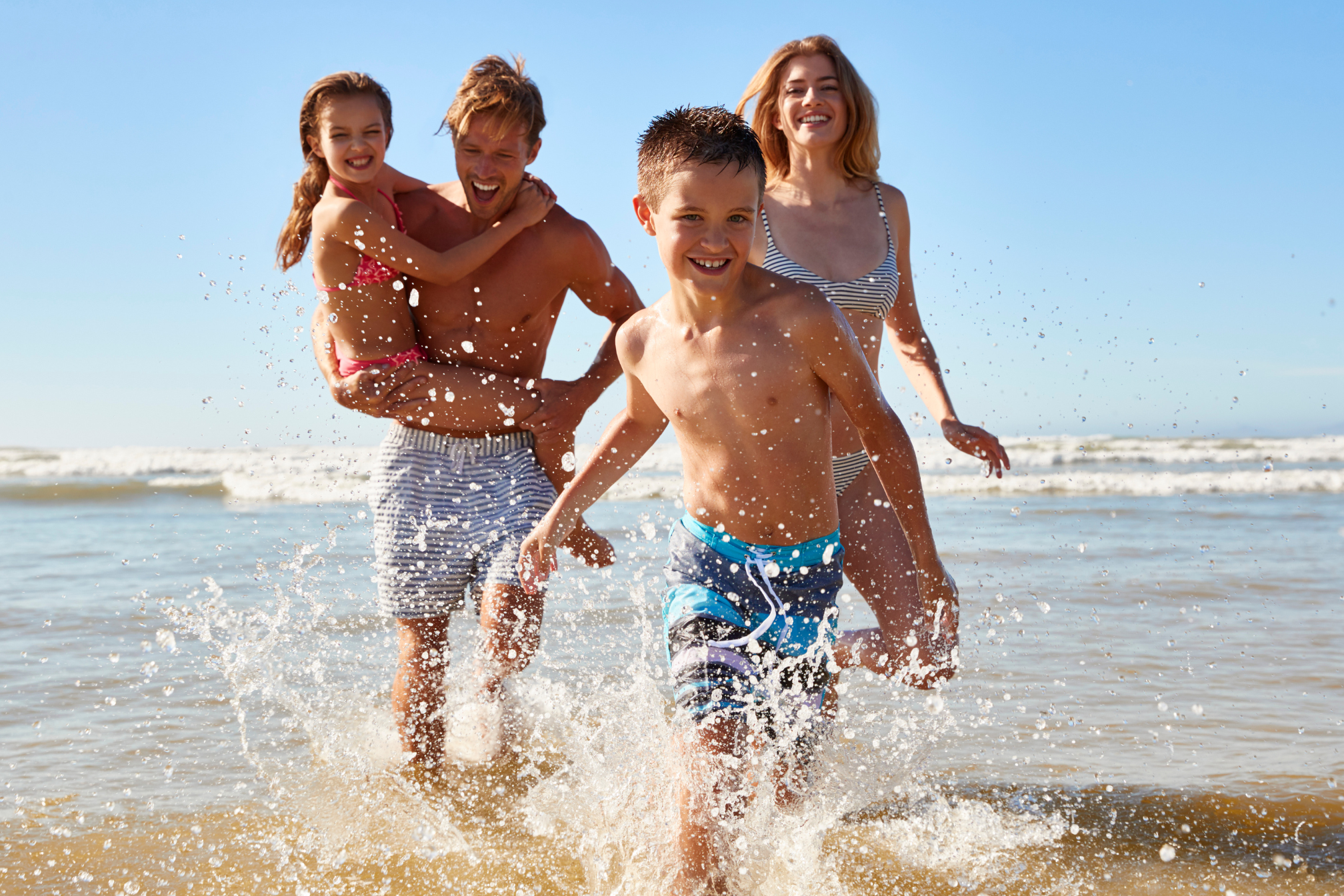 A family of four is running through the shallow water at the beach.